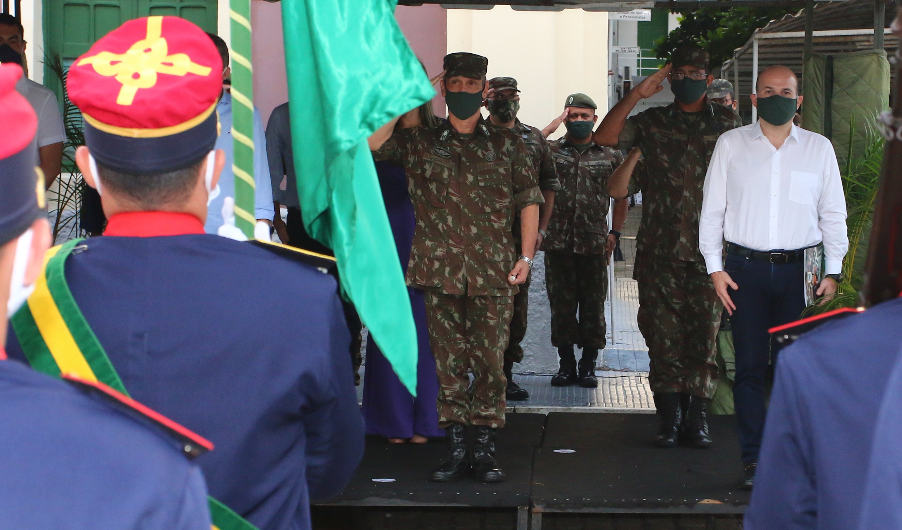 prefeito em pé ao lado de homem com roupa militar, ambos de frente para soldado que seguram bandeira